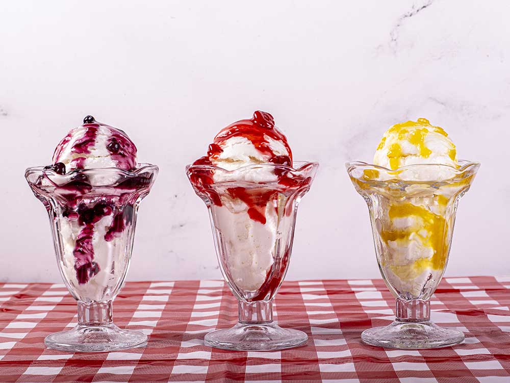 three sundaes in glasses topped with blue berry, strawberry and pineapple syrups.